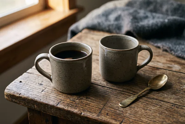 Two plain stoneware mugs resting on a weathered oak table with a brass spoon beside them, the empty second mug quietly present.
