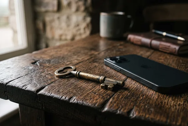 A single weathered brass key resting on an aged oak table beside a phone lying face-down.