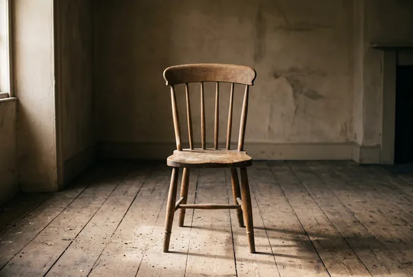 A single empty oak chair at the centre of a bare room, its worn seat and the dust on the floorboards catching the light while the rest of the room stays in soft shadow.